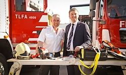 Zwei Männer stehen vor einem Feuerwehrwagen, einer trägt ein weißes Hemd und der andere einen Anzug. Sie haben Ausrüstung auf einem Tisch vor sich.