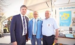 Drei Männer stehen nebeneinander vor einem Gebäude mit einem Schild, das NO Zivilschutz steht. Der Mann auf der linken Seite trägt einen Anzug und eine Krawatte, der Mann in der Mitte trägt eine blaue Jacke und der Mann auf der rechten Seite trägt ein weißes Hemd.