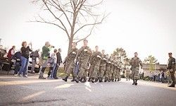 Eine Reihe von Soldaten in Tarnuniformen marschiert bei einer Parade, wobei Menschen am Straßenrand zuschauen.