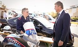 Ein Mann in einem Anzug spricht mit einem Polizeibeamten in Uniform vor einem schwarzen Motorrad und Auto, mit anderen Fahrzeugen und Personen im Hintergrund.
