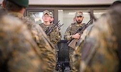 Eine Gruppe von Soldaten in Tarnkleidung, einige mit Bärten, stehen im Kreis. Sie halten Gewehre und scheinen einer Einweisung zuzuhören.