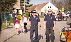 Zwei Männer in Uniform gehen die Straße entlang, während mehrere Kinder und Menschen hinter ihnen gehen. In der Ferne steht ein Gebäude und ein geparktes Fahrzeug davor.
