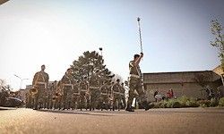 Eine Reihe von Soldaten in Uniform marschiert bei einer Parade, einer hält eine Flagge und andere spielen Musikinstrumente. Im Hintergrund steht ein Gebäude und Bäume.