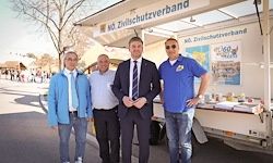 Vier Männer stehen vor einem Lastwagen mit einem Schild, das 'Keine Zivilschutzvereinigung' steht. Sie sind in formeller Kleidung gekleidet. Der Mann in der Mitte trägt eine Krawatte und der Mann auf der rechten Seite trägt eine Brille.