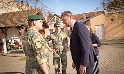 Drei Soldaten in Uniform, einer mit grüner Barett, unterhalten sich mit einem Mann im Anzug auf einer Kopfsteinpflasterstraße mit Häusern im Hintergrund.