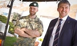 Zwei Männer stehen nebeneinander, einer in Uniform und der andere in einem Anzug. Sie lächeln beide und scheinen für ein Foto zu posieren.