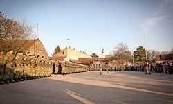 Eine Reihe von Soldaten in Uniform steht auf einer Straße, während eine Menschenmenge aus der Ferne zusieht. Im Hintergrund befinden sich Bäume und Gebäude.