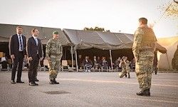 Militärangehörige in Uniform stehen in einem offenen Bereich mit Zelten. Eine Gruppe von Menschen sitzt im Hintergrund und beobachtet die Szene.