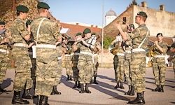 Eine Gruppe von Militärangehörigen in Tarnuniformen führt ein Musikensemble in einer Freiluftumgebung auf, mit Gebäuden im Hintergrund.