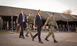 Drei Männer in Anzügen und einer in Uniform gehen auf einem gepflasterten Weg, mit Zuschauern hinter einem Zelt.