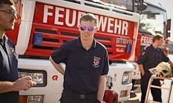 Drei Feuerwehrleute stehen vor einem Feuerwehrauto. Einer trägt eine Sonnenbrille und ein schwarzes Shirt. Die anderen beiden sind in blauen Uniformen.