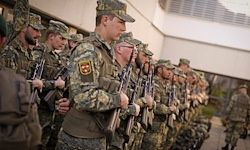 Eine Reihe von Militärangehörigen steht in Formation, hält Gewehre in den Händen und trägt Tarnuniformen mit Aufnähern auf den Schultern.
