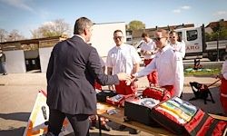 Ein Mann im Anzug schüttelt einem Mann im weißen Hemd die Hand. Sie stehen vor einem Tisch mit Erste-Hilfe-Kästen. Ein Feuerwehrauto und Menschen sind hinter ihnen.