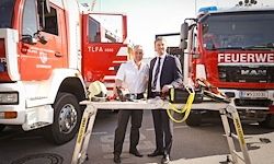 Zwei Männer stehen vor einem Tisch mit Ausrüstung vor Feuerwehrautos. Einer hält einen Helm und der andere eine Tasche. Sie tragen formelle Kleidung.