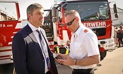 Zwei Männer in Uniformen stehen vor einem Feuerwehrauto. Einer schaut auf sein Handy, und der andere hält einen Feuerlöscher.