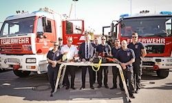 Eine Gruppe von Feuerwehrleuten steht hinter einem Tisch, wobei Feuerwehrfahrzeuge mit der Aufschrift 'Feuerwehr' im Hintergrund bei klarem Himmel zu sehen sind.