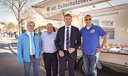 Vier Männer stehen vor einem Fahrzeug mit der Aufschrift 'No. Zivilschutzverband', einer trägt eine Sonnenbrille. Ein Mann trägt Anzug und Krawatte.