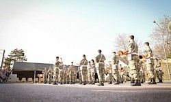 Eine Militärkapelle in Uniform steht in Formation und spielt Instrumente vor einem Gebäude, unter einem klaren blauen Himmel.