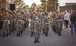 Eine Marschkapelle in Tarnuniformen spielt Musikinstrumente, während sie mit Zuschauern auf dem Bürgersteig die Straße entlanggeht.