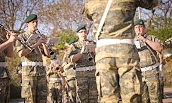 Soldaten in Tarnuniformen spielen verschiedene Musikinstrumente, darunter Trompeten, Klarinetten und Trommeln, in einer Außenumgebung mit Bäumen im Hintergrund.