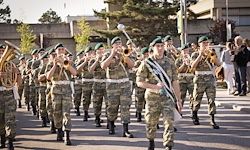 Eine Militärkapelle marschiert die Straße hinunter. Soldaten spielen Trompeten und Tuben. Sie tragen Tarnuniformen und Mützen. Die Szene ist im Freien, mit Bäumen und Gebäuden im Hintergrund.