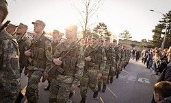 Eine Gruppe von Soldaten in Tarnuniformen marschiert mit Gewehren in der Hand eine Straße entlang, während Zuschauer von der Seite zuschauen.