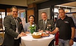 Vier Personen stehen um einen Tisch mit Essen. Zwei Männer in Uniform, einer in Anzug und einer in schwarzem Shirt. Eine Blumenvase und Bierflasche sind auf dem Tisch.