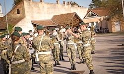 Eine Militärkapelle probt vor einem Gebäude. Die Mitglieder tragen Camouflage-Uniformen, grüne Mützen und schwarze Stiefel. Einige halten Musikinstrumente.