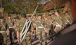 Eine Gruppe von Soldaten in Militäruniformen, einige mit Gewehr, marschiert in einer Reihe mit einem Gebäude im Hintergrund.