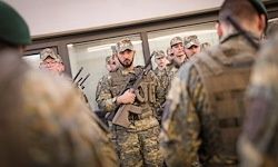 Eine Gruppe von Soldaten in Tarnuniformen steht in Formation. Ein Mann hält ein Gewehr und scheint Anweisungen zu geben.