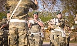 Ein Militärorchester übt im Freien mit Bäumen im Hintergrund. Die Mitglieder spielen verschiedene Instrumente, darunter Flöten und Trompeten.