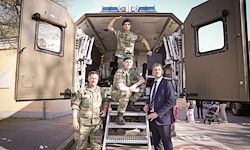 Vier Militärangehörige stehen vor einem gepanzerten Fahrzeug, einer steht auf der Treppe. Sie sind in Uniform, einer trägt einen Anzug. Im Hintergrund ist ein Gebäude und Menschen zu sehen.