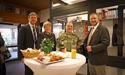 Vier Personen stehen um einen Tisch mit Essen und Getränken. Eine Frau in Uniform hält einen Blumenstrauß. Ein Mann trägt eine Krawatte.