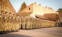 Eine Gruppe Soldaten in Tarnuniformen steht in Formation und hält Gewehre, vor einem Gebäude mit mehreren Schornsteinen und Bäumen.
