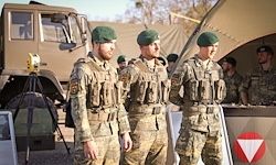 Vier Soldaten in Tarnuniformen stehen in einem militärischen Außenlager mit einem Fahrzeug und Zelten im Hintergrund. Sie tragen grüne Barette und militärische Ausrüstung.