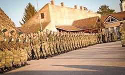 Eine Reihe von Soldaten in Militäruniformen steht in Formation vor einem Gebäude mit einer Ziegelmauer und mehreren Fenstern.