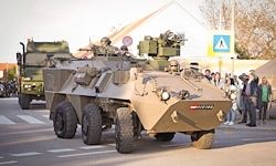 Ein gepanzertes Militärfahrzeug mit aufgesessenem Maschinengewehr und einer großen Nummer an der Seite fährt vor Schaulustigen vorüber.