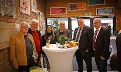Eine Gruppe von Menschen, möglicherweise Kollegen, steht um einen Tisch mit Erfrischungen und Blumen herum und posiert für ein Foto in einem Raum mit Ziegelwänden und gerahmten Bildern.