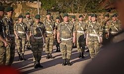 Eine Gruppe von Soldaten in Tarnuniformen steht in einer Reihe, trägt grüne Mützen, weiße Gürtel und schwarze Stiefel. Sie stehen auf der Straße, mit Bäumen und Gebäuden in der Ferne.