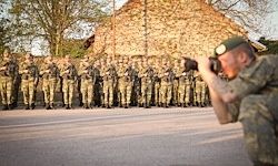 Ein Soldat in Tarnkleidung fotografiert eine Reihe von Soldaten in Uniform, die in Formation auf einer asphaltierten Straße stehen.