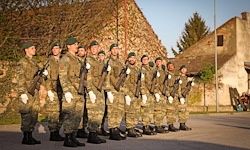 Eine Gruppe von Soldaten in Tarnuniformen, die Gewehre halten, steht in einer Reihe vor einem Steingebäude.