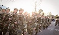 Eine Reihe von Soldaten in Tarnuniformen, die bei einer Parade marschieren. Sie tragen Gewehre und scheinen in einer militärischen Formation zu sein.