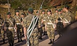 Soldaten in Militäruniformen marschieren in einer Reihe auf einer Straße, wobei eine Person vorne eine Fahne trägt. Bäume und ein Gebäude sind im Hintergrund.