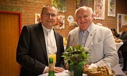 Zwei Männer sitzen an einem Tisch in einer gesellschaftlichen Umgebung. Einer trägt einen schwarzen Anzug, der andere einen weißen Blazer. Sie sind neben einer Topfpflanze und einem Korb mit Brot. Hinter ihnen befindet sich eine Ziegelwand mit hängenden Kunstwerken und einer Holztür.