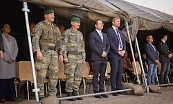 Eine Gruppe von Militärangehörigen und Beamten steht unter einem Zelt und beobachtet eine formelle Veranstaltung. Sie tragen Uniformen und Anzüge.