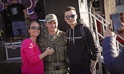 Ein Mann in Militäruniform und eine Frau in Pink stehen nebeneinander, mit einem Mann in einer schwarzen Kapuzenjacke neben ihnen, alle lächeln für ein Foto.