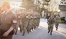 Eine Gruppe von Soldaten in Uniform marschiert auf der Straße, mit Zuschauern im Hintergrund. Sie tragen Gewehre und Handschuhe.