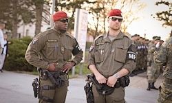 Zwei Militäroffiziere stehen im Freien. Beide tragen Sonnenbrillen und haben Waffen bei sich. Eine Reihe anderer Soldaten steht hinter ihnen.