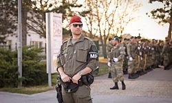 Ein Soldat in einem roten Barett steht vor einer Reihe von Soldaten in Uniform. Sie stehen alle draußen auf einem Betonweg.