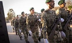 Eine Gruppe von Soldaten in Militäruniformen und grünen Mützen marschiert die Straße entlang, trägt Gewehre und trägt weiße Handschuhe.